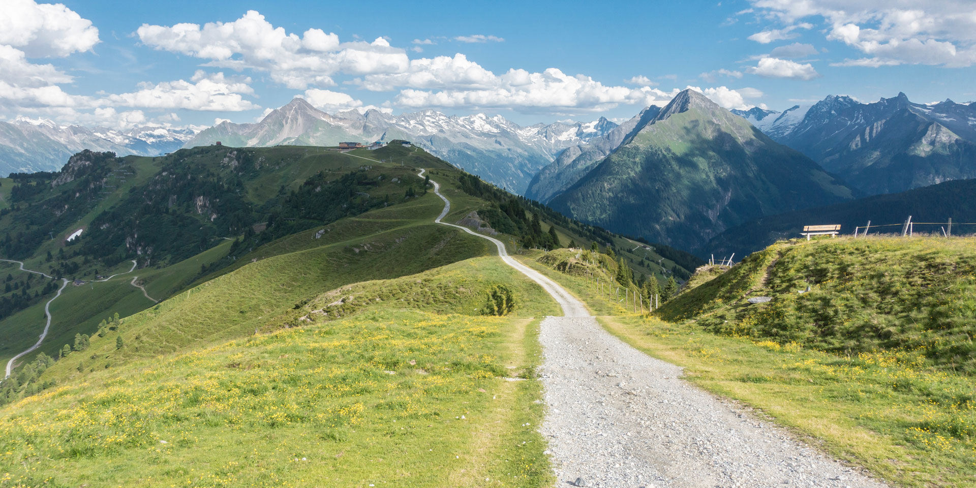 Eine wunderschöne alpine Landschaft mit einem gewundenen Schotterweg, der durch grüne Wiesen führt, während hohe Berge und schneebedeckte Gipfel in der Ferne zu sehen sind. Eine Bank steht auf dem Hügel und bietet einen malerischen Ausblick.