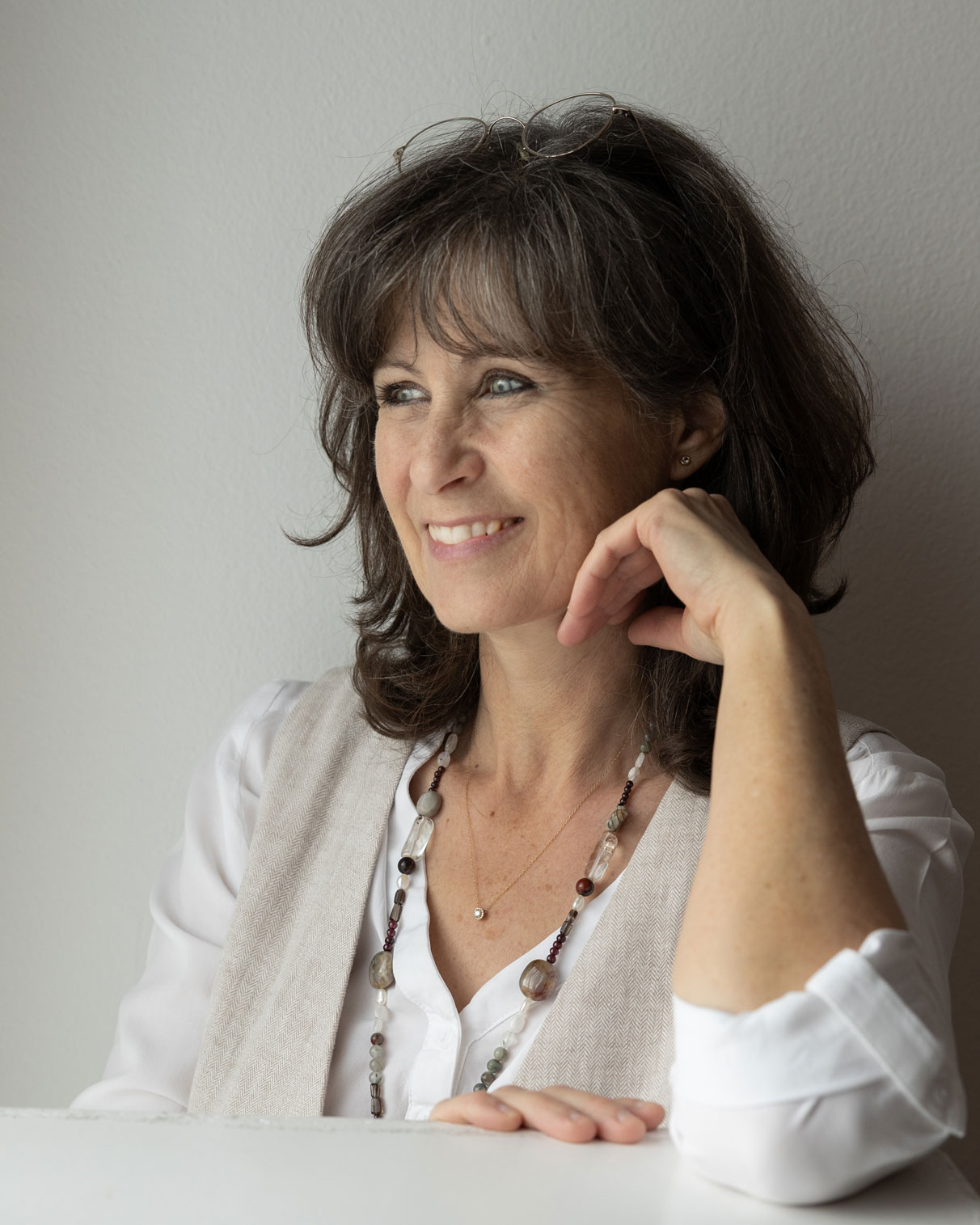 Ein nachdenkliches Portrait von Andrea Engel, die ein weisses Shirt und eine beigen Weste trägt. Sie lächelt sanft, während sie ihr Kinn auf die Hand stützt, und eine Perlenkette ist um ihren Hals zu sehen. Der sanfte, neutrale Hintergrund hebt ihre ruhige und nachdenkliche Ausstrahlung hervor.