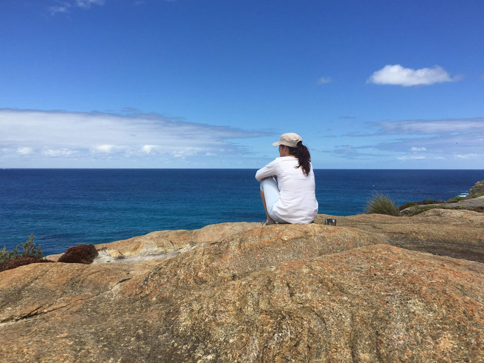 Eine Person, die auf einem grossen Felsen am Meer sitzt, ein weisses Shirt und eine Kappe trägt und auf das blaue Wasser unter einem klaren Himmel blickt. Die Szene vermittelt einen ruhigen Moment der Reflexion und Verbindung mit der Natur.