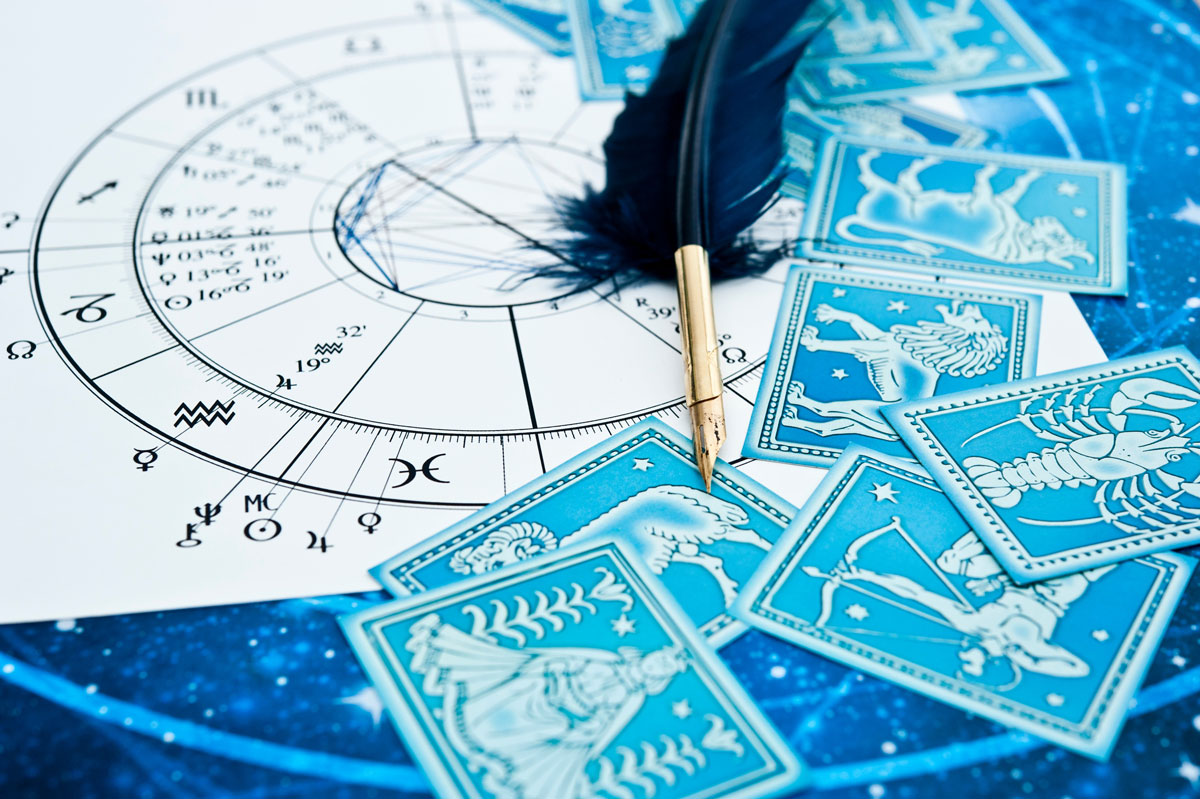 Eine Nahaufnahme einer astrologischen Karte mit Tierkreiszeichen und einem Federkiel. Mehrere Tierkreis-Karten, die verschiedene astrologische Symbole zeigen, sind über den Tisch verteilt, wobei der kosmische blaue Hintergrund das mystische Thema unterstreicht.