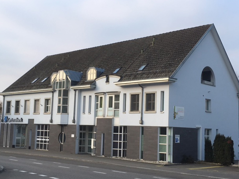 Ein modernes weisses Bürogebäude mit grossen Fenstern, einem geneigten Dach und einer Bank auf der linken Seite des Bildes. Das Gebäude befindet sich an einer ruhigen Strasse mit einer kleinen grünen Fläche auf der rechten Seite.