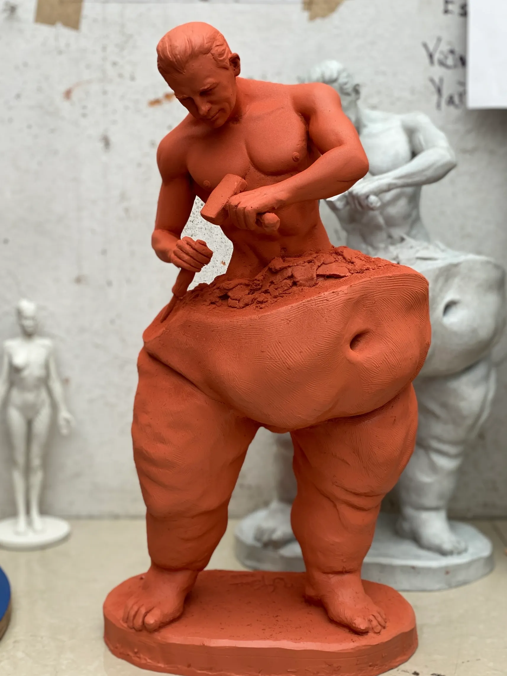 Eine Skulptur in Arbeit, die einen muskulösen Mann zeigt, der an einem Block rotem Ton meisselt. Die Figur ist unvollständig, mit einer groben Textur und einer kraftvollen Pose, vor einem neutralen Hintergrund mit anderen Skulpturen.